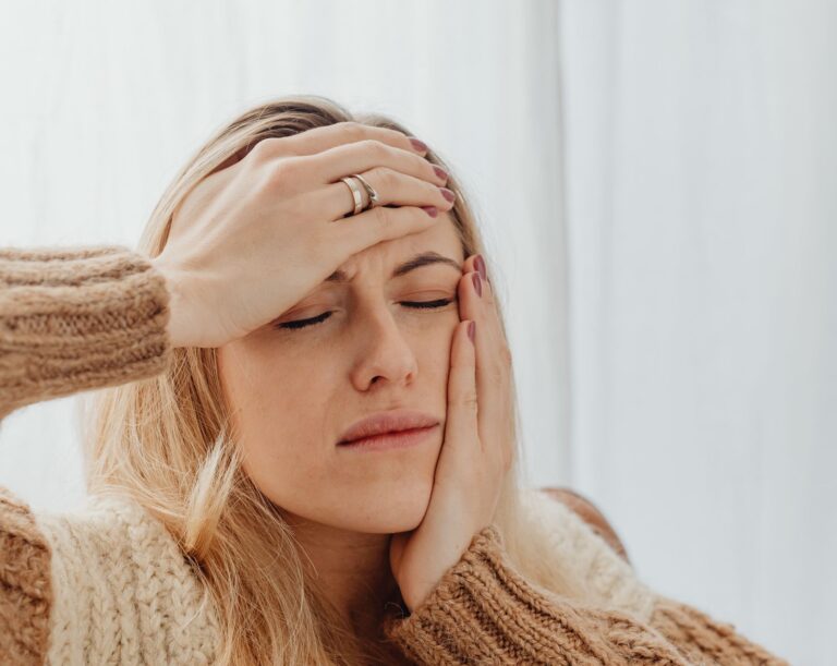 Woman with closed eyes holding head, wearing a knitted sweater, possibly experiencing discomfort.